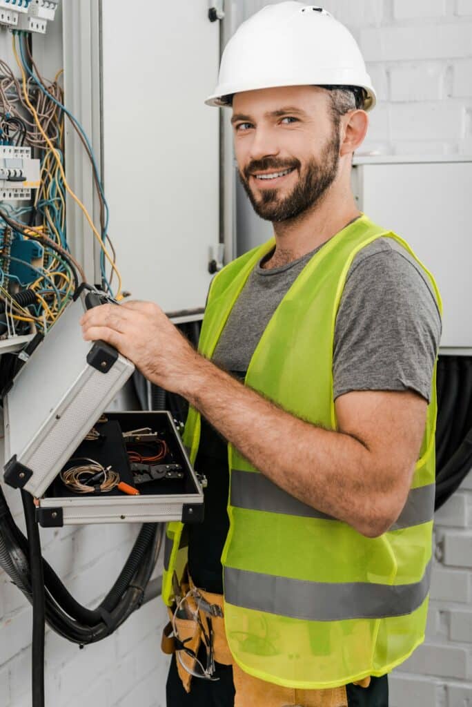 smiling handsome electrician holding toolbox near electrical box in corridor and looking at camera